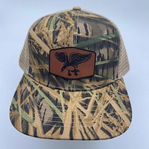 May include: A camouflage baseball cap featuring a tan mesh back and a brown leather-like patch on the front. The patch is shaped like a hexagon with rounded corners and displays a black graphic of a flying duck in the center of the design.