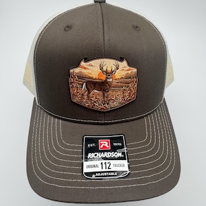 May include: A brown and white trucker hat featuring a patch with a deer standing in a field at sunset. The front of the hat displays a black sticker with the text EST. 1970 RICHARDSON ORIGINAL 112 TRUCKER ADJUSTABLE and a red R logo.