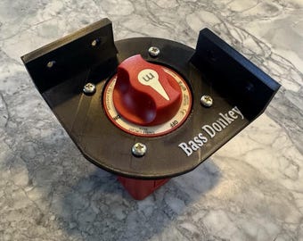 May include: A black plastic switch with a red knob and the words "SINGLE CIRCUIT 600A" and "BLUE SEA SYSTEMS" printed on it. The switch is mounted on a black plastic base with the words "Bass Donkey" printed on it.