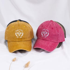May include: Several baseball caps in shades of mustard yellow and fuchsia. Each cap has embroidered text and a tennis-themed emblem. The mustard cap says "FORSTER EST 1984". The fuchsia cap reads "ADELAIDE EST 1996".