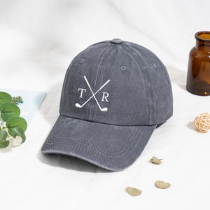 Könnte beinhalten: Graue Baseballkappe mit gebogenem Schirm. Die Kappe hat eine weiße Grafik mit gekreuzten Golfschlägern und den Buchstaben "T" und "R". Die Kappe ist aus einem weichen Material gefertigt.