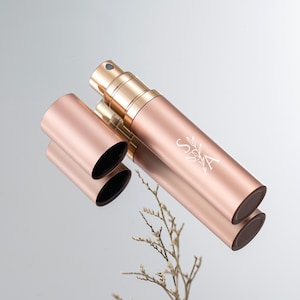 May include: A rose gold perfume atomizer with a matching cap, on a reflective surface. The atomizer has a gold spray nozzle and the letters "S&A" with a floral design. The cap is detached, revealing a black interior.