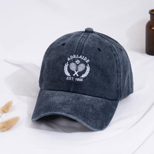 May include: A dark grey baseball cap with a curved peak. The cap has white embroidered text reading "ADELAIDE" above a tennis racket design and "EST. 1996". The cap is made of a soft, worn material.
