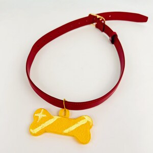 Può includere: Un collare rosso con una fibbia in metallo dorato e una targhetta gialla a forma di osso. La targhetta presenta una superficie testurizzata con due strisce diagonali giallo chiaro e un piccolo dettaglio a forma di stella.