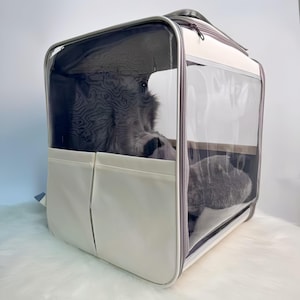 May include: A cream-colored pet carrier with clear plastic and mesh panels. The carrier has a zippered top and two front pockets. A gray cat is visible inside the carrier.