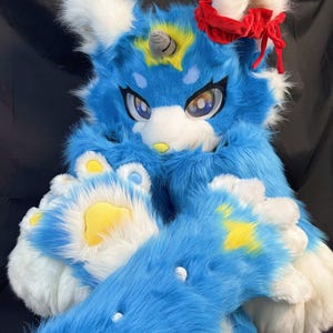 May include: A blue and white furry fursuit with a unicorn horn and a red bow. The fursuit has large eyes, yellow accents, and paw-like hands and feet. The overall design is cartoonish and playful.