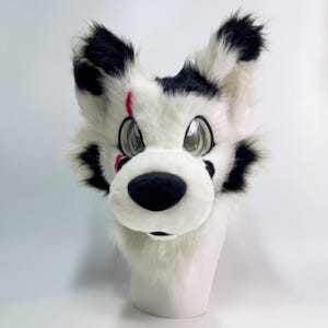 May include: A white and black furry animal head with large, expressive green eyes and a black nose. The ears and cheeks have black spots. A red scar is visible on the left side of the face. The head is mounted on a white mannequin head.