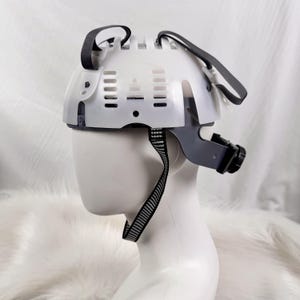 May include: White safety helmet with black straps and a grey adjustable chin strap. The helmet has a vented design with multiple holes.