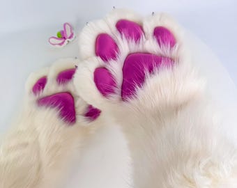 Puede incluir: Un par de guantes de pata de piel blanca con almohadillas de pata de color rosa brillante. Una pequeña decoración de mariposa rosa y verde es visible. Los guantes están diseñados para parecerse a patas de animales.