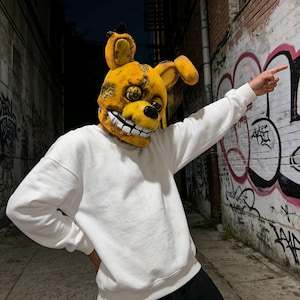 May include: A yellow and brown Springtrap mask with large ears and a wide, toothy grin. The mask is worn with a white sweatshirt and black pants. The background is a dark alley with graffiti-covered walls.