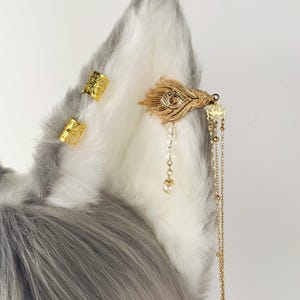 May include: A faux fur ear with gold-colored decorative accents. The ear has a gray and white color scheme. The gold accents include small rectangular pieces and a feather-shaped piece with dangling chains and beads.