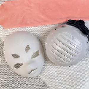Può includere: Sono mostrate due maschere bianche. Una è un volto vuoto con ritagli per occhi e bocca. L'altra è una maschera bianca ventilata con un cinturino nero. Un tessuto rosa è sullo sfondo.
