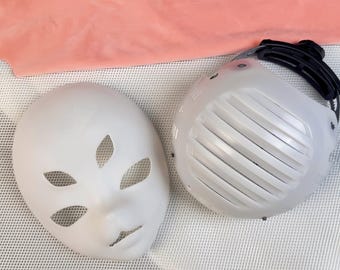 May include: Two white masks are displayed. One is a blank face with eye and mouth cutouts. The other is a white, vented mask with a black strap. A pink fabric is in the background.