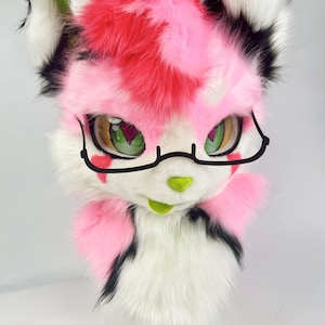 Puede incluir: Una cabeza de animal peluda rosa, blanca y negra, con grandes ojos verdes y gafas negras. La cabeza tiene una nariz rosa y una boca verde. El pelaje es una mezcla de rosa, blanco y negro, creando una apariencia juguetona y caprichosa.
