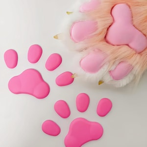 May include: A close-up of a pink and white furry paw with pink paw pads and yellow claws. Two sets of pink paw prints are on a white background. The paw pads are a vibrant pink color.