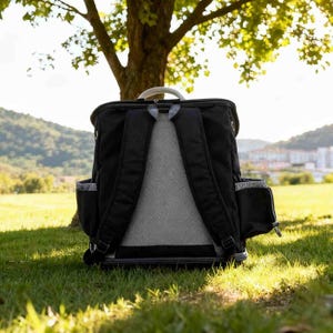 K&ouml;nnte beinhalten: Schwarzer Rucksack mit gepolstertem R&uuml;cken und Schultergurten, auf gr&uuml;nem Gras. Der Rucksack hat Seitentaschen und einen oberen Griff. Der Hintergrund zeigt einen Baum und eine Aussicht auf eine entfernte Stadt.