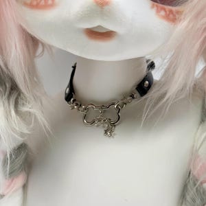 May include: A black leather choker necklace with a silver-colored bone-shaped clasp and star charms. The necklace is worn on a white mannequin with pink and gray hair.