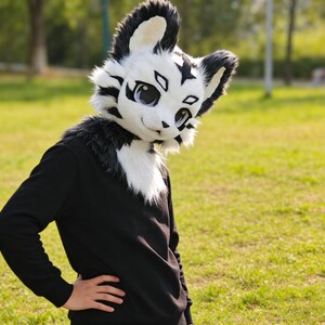 May include: A white and black furry animal mask with large eyes and a smiling expression. The mask has black accents and is worn with a black long-sleeve shirt. The background is a grassy field.