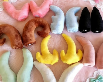 以下が含まれることがあります： ピンク、茶色、青、黒、黄色、緑、白など、さまざまな色のぬいぐるみホーンのコレクション。 ホーンはピンク色のふわふわした表面に配置され、縁には装飾的なリボンが付いています。