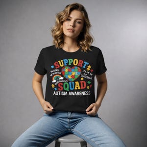 Puede incluir: Camiseta negra con un gráfico colorido que apoya la concienciación sobre el autismo. El diseño incluye un rompecabezas en forma de corazón, un arcoíris y las palabras "SUPPORT", "SQUAD" y "AUTISM AWARENESS". La camiseta también tiene el texto "In April We Wear Blue" y "It's ok to be different."