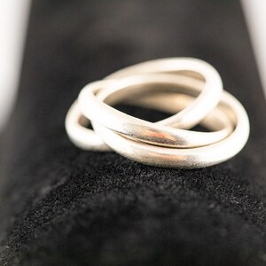 May include: A silver-toned triple band ring featuring three interlocking smooth, rounded metal bands that overlap to form a single cohesive piece of jewellery with a polished, reflective surface finish and a continuous circular structure.