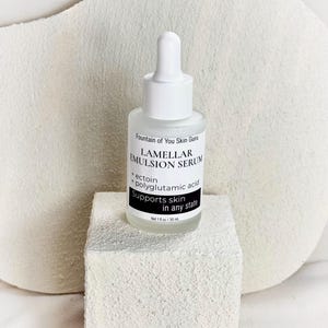 May include: A white glass bottle of Lamellar Emulsion Serum with a dropper top, resting on a white textured block. The label reads "Fountain of You Skin Guru" and lists ingredients. The bottle contains 1 fl oz / 30 mL of product.