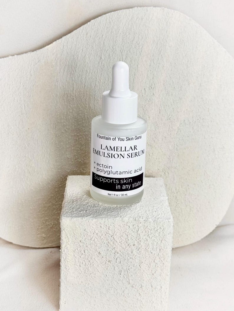 May include: A white glass bottle of Lamellar Emulsion Serum with a dropper top, resting on a white textured block. The label reads "Fountain of You Skin Guru" and lists ingredients. The bottle contains 1 fl oz / 30 mL of product.