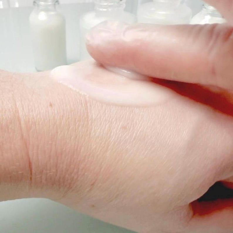 May include: A close-up of a hand applying a white, viscous liquid. The skin is light-toned, and the liquid appears to be a lotion or cream. Several small glass bottles with white liquid are visible in the background.