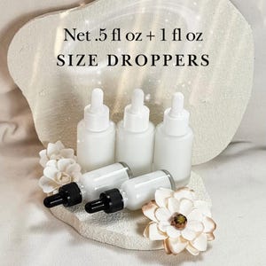 May include: White glass dropper bottles with black droppers are arranged on a textured surface, accompanied by white and cream-colored floral accents. The text "Net .5 fl oz + 1 fl oz SIZE DROPPERS" is displayed at the top.