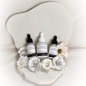 May include: Three white skincare serum bottles with black droppers are arranged on a textured, off-white decorative base, adorned with paper flowers. The bottles are labeled "Lamellar Emulsion Serum" with text that reads "ectoin + polyglutamic acid Supports skin in any state."