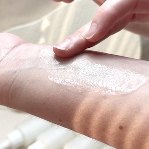 May include: A hand applying a white, creamy substance to the wrist. The substance is spread across the skin. Several white pump bottles are visible in the background, suggesting a skincare product.