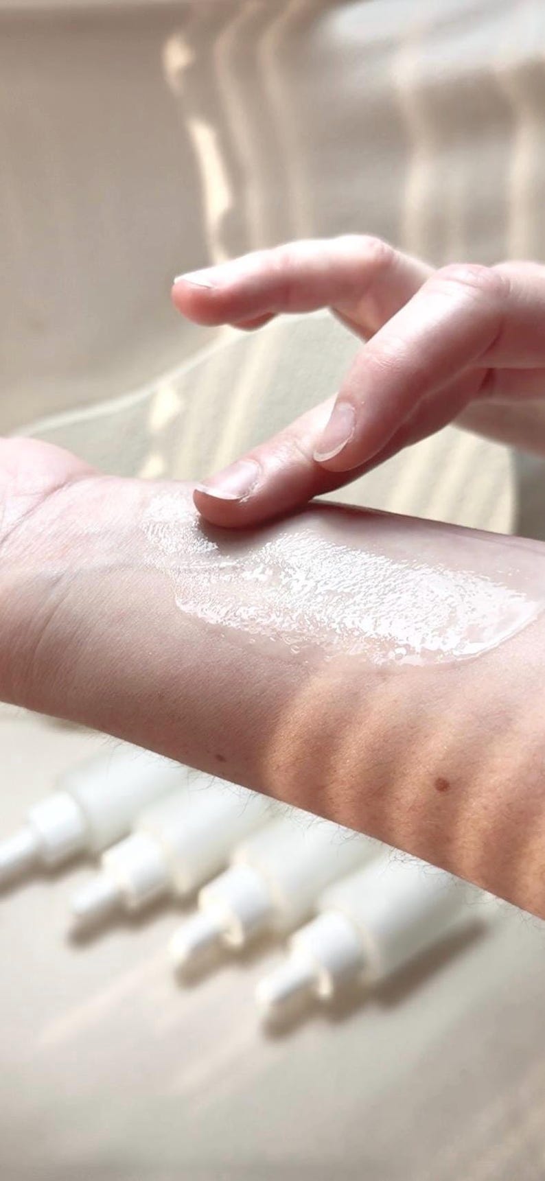 May include: A hand applying a white, creamy substance to the wrist. The substance is spread across the skin. Several white pump bottles are visible in the background, suggesting a skincare product.