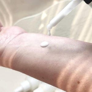 May include: A close-up of a white dropper dispensing a white liquid onto a person's arm. A small, round drop of the liquid sits on the skin. The dropper is held at an angle, with a single drop falling.