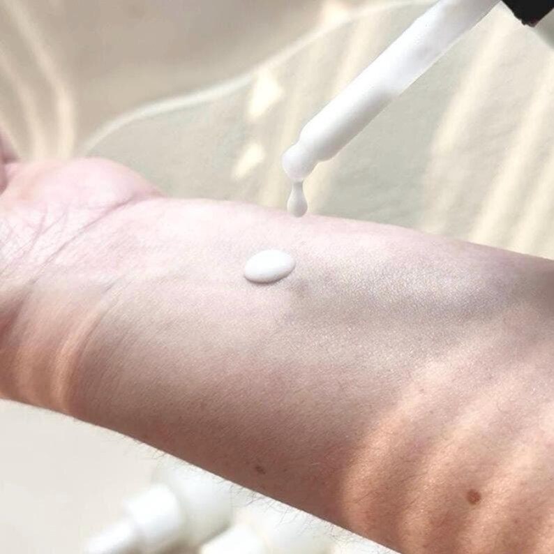 May include: A close-up of a white dropper dispensing a white liquid onto a person's arm. A small, round drop of the liquid sits on the skin. The dropper is held at an angle, with a single drop falling.