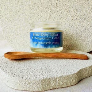 Puede incluir: Un tarro de cristal de Bone-Deep Balm X2 Magnesium Conc. con una etiqueta azul y blanca, y una cuchara de madera. La etiqueta dice "quiet limbs &bull; settle tension". El tarro est&aacute; sobre una superficie blanca y texturizada.