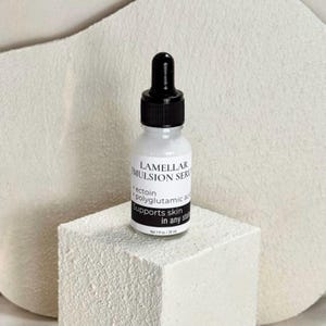 May include: A white bottle of Lamellar Emulsion Serum with a black dropper top. The label includes the text "ectoin + polyglutamic acid Supports skin in any state". The bottle is on a white textured block.