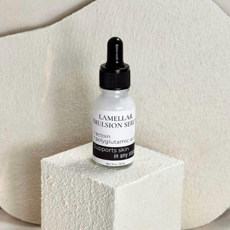 May include: A white bottle of Lamellar Emulsion Serum with a black dropper top. The label includes the text "ectoin + polyglutamic acid Supports skin in any state". The bottle is on a white textured block.