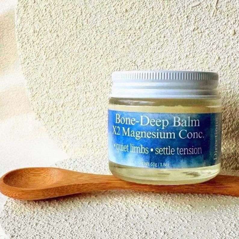 Puede incluir: Un tarro de cristal de Bone-Deep Balm X2 Magnesium Conc. con tapa blanca, sobre una cuchara de madera. La etiqueta dice "quiet limbs &bull; settle tension." El tarro contiene 51g de b&aacute;lsamo.