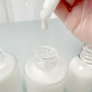 May include: Close-up of white liquid dripping from a dropper into a clear glass bottle. The bottle is surrounded by two other bottles. The background is a light, neutral color.