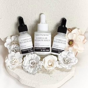 May include: Three white serum bottles with black droppers are arranged with paper flowers. The bottles are labeled "Lamellar Emulsion Serum" and "Fountain of You Skin Guru." The text on the bottles includes "ectoin," "polyglutamic acid," and "Supports skin in any state."