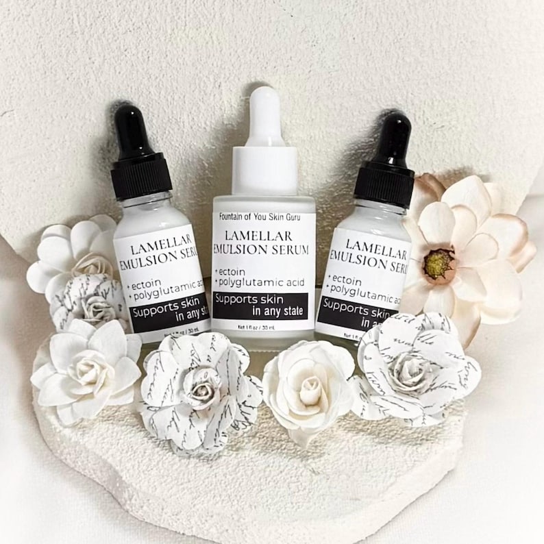 May include: Three white serum bottles with black droppers are arranged with paper flowers. The bottles are labeled "Lamellar Emulsion Serum" and "Fountain of You Skin Guru." The text on the bottles includes "ectoin," "polyglutamic acid," and "Supports skin in any state."