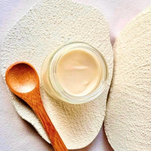 Puede incluir: Un tarro de crema de color beige claro se encuentra sobre una superficie texturizada de color blanquecino. Una cuchara de madera descansa junto al tarro. La crema est&aacute; en un tarro de vidrio transparente, y la cuchara es de madera.