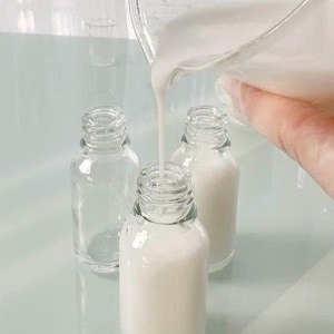 May include: Clear glass bottles being filled with a white liquid. One bottle is empty, while the others are partially or fully filled. The liquid is being poured from a clear container, suggesting a cosmetic or skincare product.