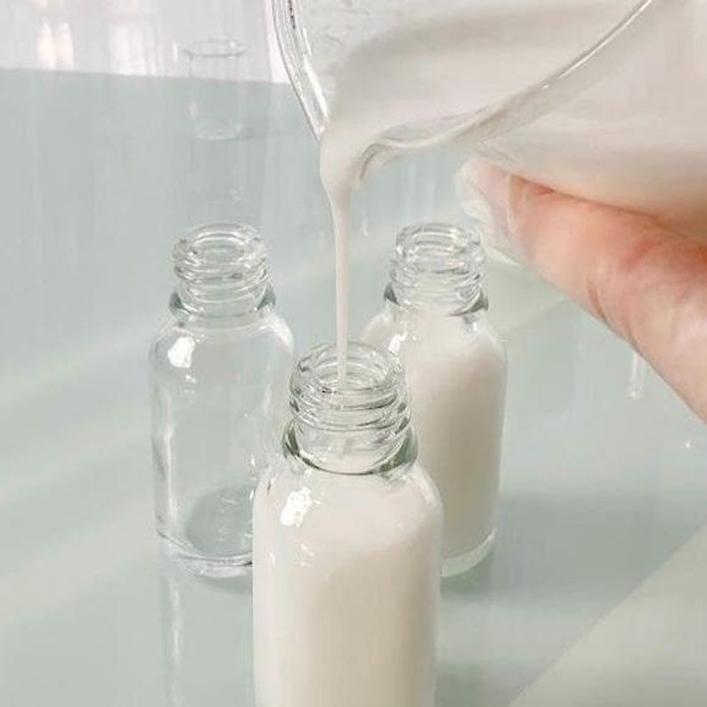 May include: Clear glass bottles being filled with a white liquid. One bottle is empty, while the others are partially or fully filled. The liquid is being poured from a clear container, suggesting a cosmetic or skincare product.