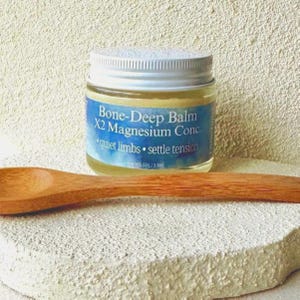 Puede incluir: Un peque&ntilde;o frasco de vidrio de Bone-Deep Balm X2 Magnesium Conc. con tapa plateada y etiqueta azul. La etiqueta dice "quiet limbs &bull; settle tension". Una cuchara de madera descansa junto al frasco sobre una superficie blanca texturizada.