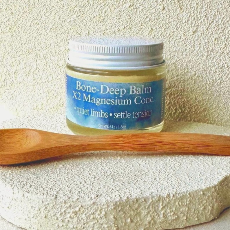 Puede incluir: Un peque&ntilde;o frasco de vidrio de Bone-Deep Balm X2 Magnesium Conc. con tapa plateada y etiqueta azul. La etiqueta dice "quiet limbs &bull; settle tension". Una cuchara de madera descansa junto al frasco sobre una superficie blanca texturizada.