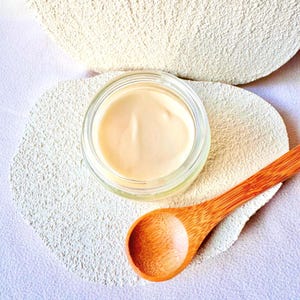 Puede incluir: Un peque&ntilde;o tarro de vidrio transparente lleno de una sustancia cremosa de color beige claro. Una cuchara de madera descansa junto al tarro sobre una superficie texturizada de color blanquecino. El fondo presenta m&aacute;s del mismo material.