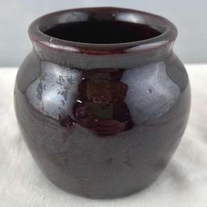 Könnte beinhalten: Ein kleiner, dunkelbrauner Keramiktopf mit glänzender Oberfläche. Der Topf hat einen abgerundeten Körper und einen breiten, leicht ausgestellten Rand. Das Innere ist dunkel, und die Außenseite reflektiert das Licht und zeigt subtile Farbvariationen.