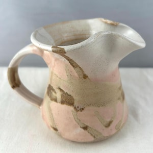 May include: A ceramic pitcher with a handle and a ruffled spout. The pitcher features a speckled, off-white rim and handle, with a pink and tan pattern on the body. The pitcher is likely handmade and suitable for serving beverages.