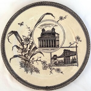 May include: Cream-coloured decorative plate with a black and white floral and architectural design. Features illustrations of the Pantheon in Rome and the Brooklyn Bridge. The plate has a detailed border.